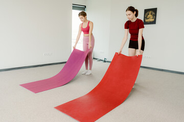 Two young women are setting up colorful yoga mats in a spacious studio. They share smiles while...
