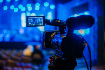 Professional video camera positioned on tripod, capturing live performance stage with vibrant blue lighting, showcasing the art of videography and event documentation