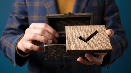 A person in a stylish plaid blazer carefully holds an opened rustic wooden box, showcasing its unique lid which features a prominent checkmark symbol expertly cut from coarse, textured burlap. The det