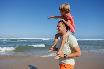 Child boy on dad's shoulders, pointing at sea under clear sky