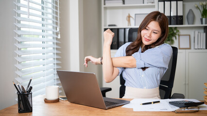 Businesswoman stretching her arm while sitting at a desk in front of a laptop.