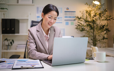 Smiling businesswoman working on a laptop in a bright, modern office with documents and coffee on the desk.
