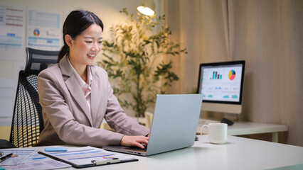 Confident businesswoman working on laptop in a modern office setting with documents and analytics in the background.