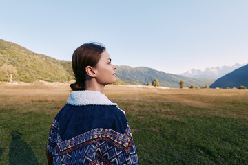 Woman in patterned jacket stands outdoors in a meadow, profile view facing mountains and expansive landscape. Nature travel scene with serene sky and calm countryside mood. © SHOTPRIME STUDIO