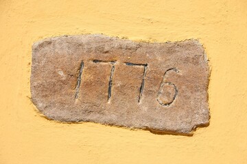 Close-up of an old stone plaque with a 4-digit house number hanging on a yellow historic house wall