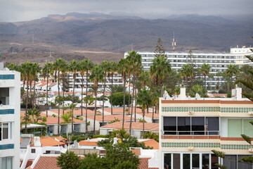 Multi-story buildings, residential, hotel complexes against Gran Canaria's mountains. Buildings have architectural style with white facades, tiled roofs. Palm trees are visible throughout the scene. 