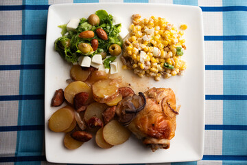 Healthy Mediterranean Dinner with Green Salad, Olives, and Corn Salad, Roasted Chicken Thigh with Sliced Potatoes and Sausage on a White Plate