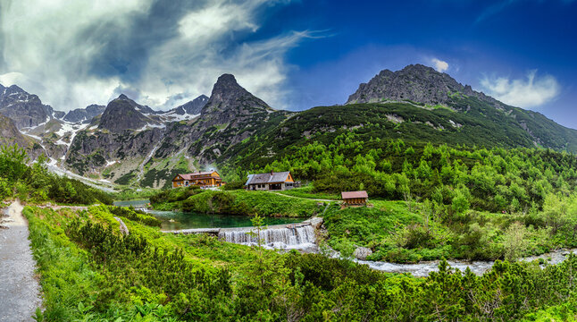 A wide panoramic view of the hiking area at Zelene pleso. The image shows the main mountain hut, smaller service buildings, a stone dam with a waterfall, and a hiking path, all set against a dramatic 