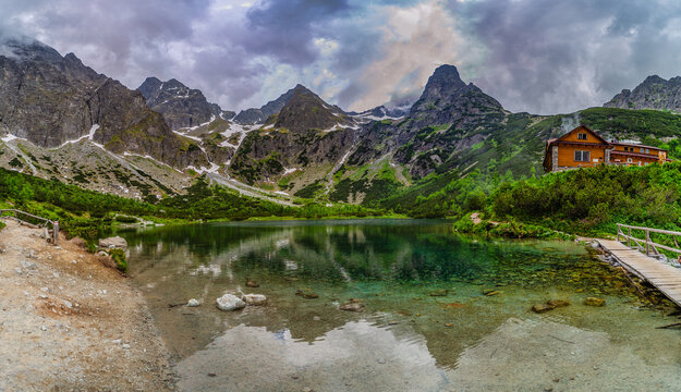 A dramatic landscape shot of a hiking path at the edge of Green Lake (Zelene pleso). Massive granite peaks with snow patches are shrouded in heavy grey storm clouds. Tatra National Park, Slovakia.