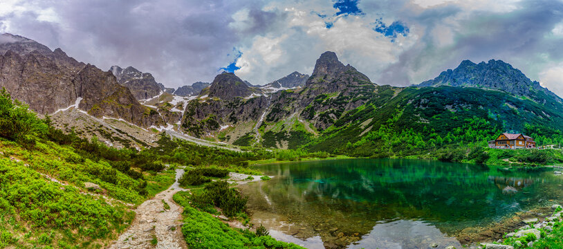 A dramatic landscape shot of a hiking path at the edge of Green Lake (Zelene pleso). Massive granite peaks with snow patches are shrouded in heavy grey storm clouds. Tatra National Park, Slovakia.