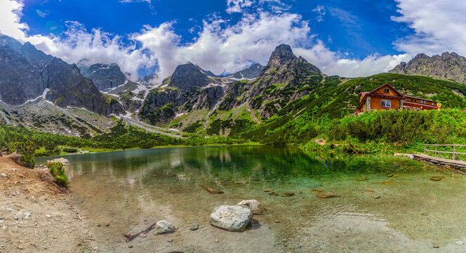 A breathtaking wide panorama of Zelene pleso (Green Lake) in the High Tatras, Slovakia. The image features the iconic mountain hut, crystal clear water with a rocky bottom, and jagged granite peaks un
