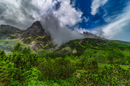 A scenic view of the mountain cabin "Chata pri Zelenom plese" nestled in a lush green valley. Surrounded by high rocky peaks and dark, moody storm clouds in the High Tatras National Park, Slovakia.