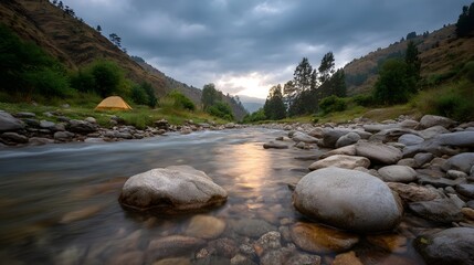 A scenic river flows through a valley at golden hour with a yellow tent set up on the bank under cloudy skies
