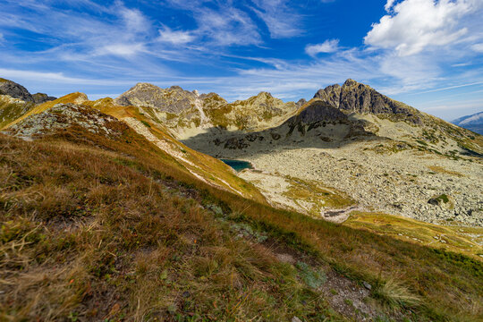 A narrow rocky path winds up a steep grassy slope toward a mountain summit. Scenic view from Hladke sedlo in High Tatras National Park, featuring a turquoise lake in the valley under a cloudy blue sky