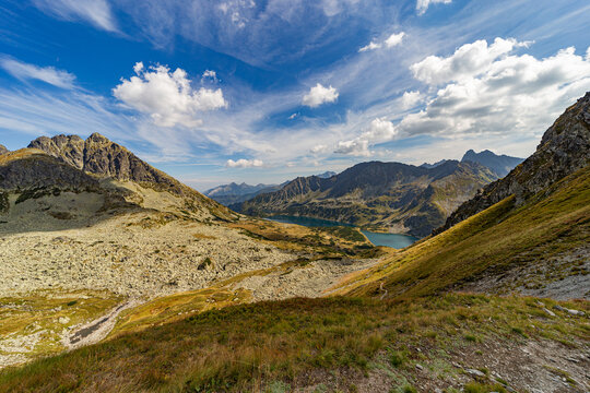 A narrow rocky path winds up a steep grassy slope toward a mountain summit. Scenic view from Hladke sedlo in High Tatras National Park, featuring a turquoise lake in the valley under a cloudy blue sky