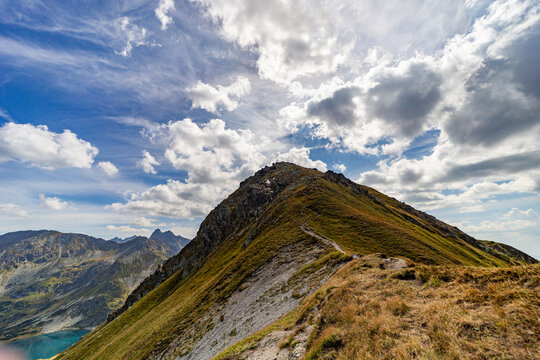 A narrow rocky path winds up a steep grassy slope toward a mountain summit. Scenic view from Hladke sedlo in High Tatras National Park, featuring a turquoise lake in the valley under a cloudy blue sky