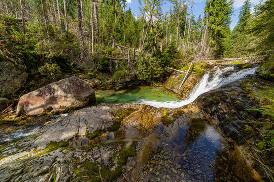 A beautiful waterfall flows into a crystal clear emerald pool in a lush green forest. Captured in the High Tatras National Park, Slovakia, showcasing pristine alpine nature and sunlit rocks.