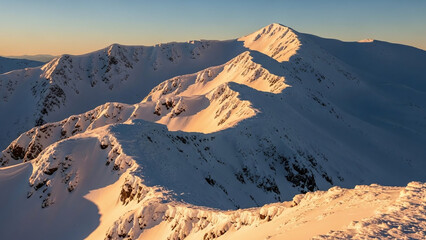 Breathtaking snowy mountain range at sunrise, golden light on peaks creating serene atmosphere with rugged terrain and clear blue sky