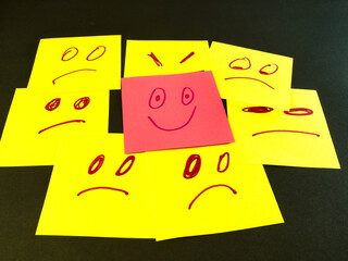 Happy Face Amidst Sad Faces - Red happy face sticky note stands out from numerous yellow sad and angry faces on a dark background, symbolizing positivity and uniqueness.