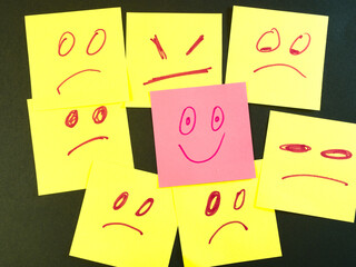 Happy Face Stands Out in Crowd of Sad - One pink sticky note with a happy face stands out from many yellow sticky notes displaying sad and angry expressions. Uniqueness.