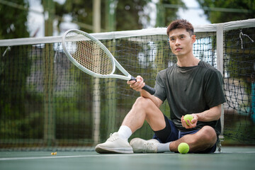 Athletic man sitting on a tennis court, holding a tennis ball while taking a thoughtful pause during training.