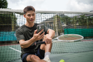 Young man sitting on a tennis court, smiling while checking his phone during a break from practice