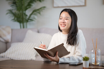 Young woman in a white sweater journaling at a wooden coffee table in a bright, modern living room.