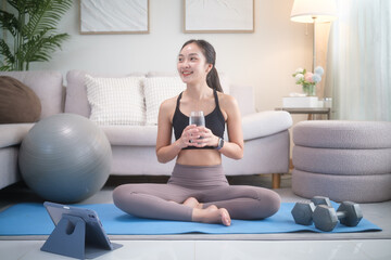 Smiling woman sitting on a yoga mat at home, drinking water after an exercise session. Home workout, hydration, and healthy lifestyle concept.