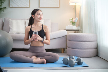 Young woman holding a glass of water, relaxing after exercise on a yoga mat. Healthy lifestyle,...