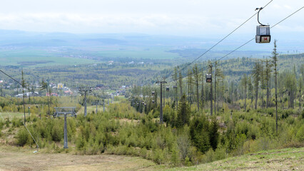 Cable car gondola ascending mountain in Tatranska Lomnica ski resort, Slovakia. Scenic summer view with green forests, alpine slopes, and distant valley landscape under cloudy sky. © Arseniy
