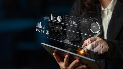 A professional woman interacts with a tablet displaying advanced data visualization graphics, showcasing analytics and insights in a dark office environment. Scalp
