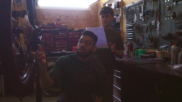 Two young male mechanics collaborating on a bicycle repair, with one reading instructions and the other examining the bike before they select the right tools from a pegboard in their workshop
