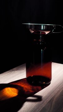 Hand placing a stainless steel funnel into the opening of an empty amber glass jar. Dark moody lighting with caustic reflections. Preparation for refilling pantry ingredients.