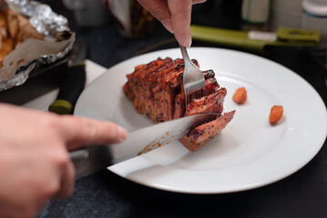 Juicy pork roast is being sliced on a clean white plate. Hands hold knife and fork, with foil-wrapped meat in the background, conveying a warm home kitchen meal