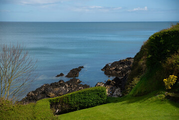 A serene coastal scene with a rocky shoreline, lush green grass, and calm blue sea beneath clear sky. View of the Atlantic Ocean from the green island of Ireland