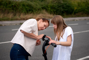 Two friends inspect and adjust an electric scooter on a suburban roadside, sharing moment of focus and collaboration. Young girl teaches her mother how to setup an electric scooter