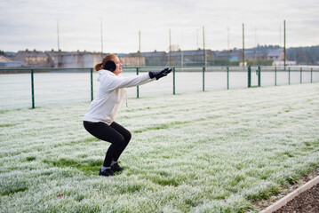 A fit woman in a white hoodie and black leggings performs a squat in frost-covered field beside a fence, outdoor fitness, cold weather training, and determination