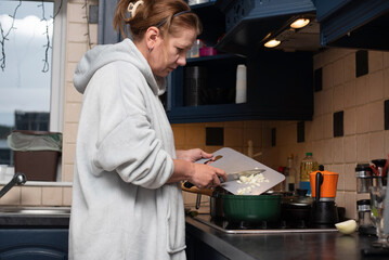 A woman in a cozy robe chops onions on a cutting board over a stove, preparing meal in a modern kitchen. A woman prepares meat gravy at home, adding lots of onions, carrots, and meat