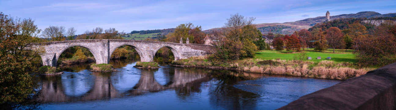 Stirling Old Bridge spanning the River Forth with the National Wallace Monument rising beyond, a tranquil and vibrant autumn landscape and the landmark historic architectures of Scotland in UK