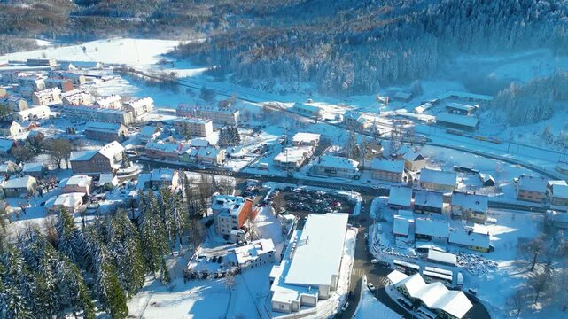An aerial view of Delnice, Croatia, blanketed in pristine winter snow. A charming town nestled amidst forested hills under a clear blue sky.