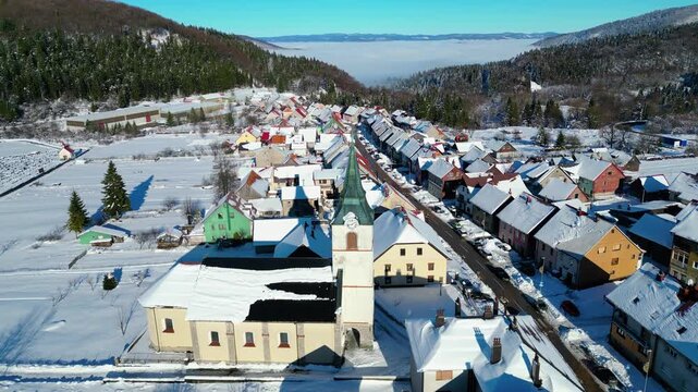 Winter's embrace in a charming Croatian village Delnice, with snow-dusted roofs and a prominent church under a clear blue sky. Fog blankets the distant valley.