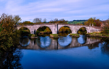 Fototapeta premium Stirling Old Bridge spanning the River Forth with the National Wallace Monument rising beyond, a tranquil and vibrant autumn landscape and the landmark historic architectures of Scotland in UK