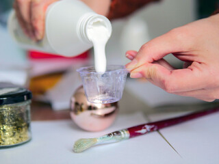 Hands Pouring White Glue into a Small Mixing Cup for Crafts - DIY Holiday Crafting: Pouring Liquid Adhesive with Glitter and Paintbrushes on a Desk