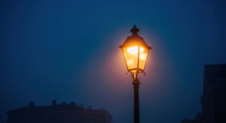 A solitary illuminated street lamp casts a warm glow against a dark, foggy blue sky