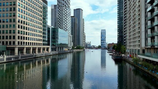 Aerial pull back shot along the docks of Canary Wharf, London. Modern waterfront cityscape with office and residential buildings, calm water reflections and urban life. Reverse dolly shot.