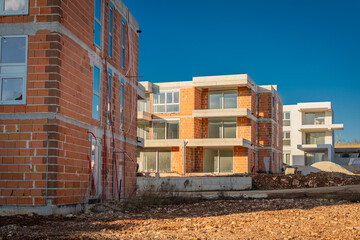 A modern residential complex under construction features multi-story buildings with exposed red clay bricks, concrete slabs, and installed glass windows against a clear, deep blue horizon.