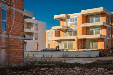 A modern residential complex under construction features multi-story buildings with exposed red clay bricks, concrete slabs, and installed glass windows against a clear, deep blue horizon.