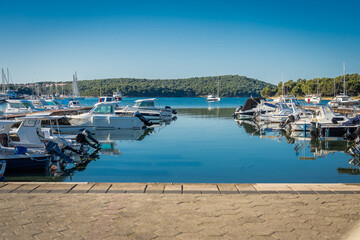 A peaceful marina in Medulin, Croatia, features various motorboats moored in calm, turquoise waters. Lush green hills and a clear blue sky provide a serene backdrop for this coastal scene.