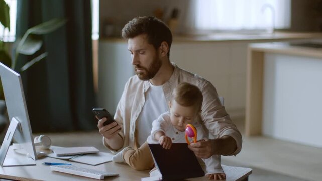 In a cozy kitchen, a father multitasks by checking his phone while his young baby plays with a toy on a desk. Natural light highlights their warm interaction.
