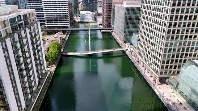 Aerial backward dolly shot over the docks of Canary Wharf, London. Modern waterfront cityscape with office buildings, pedestrian bridge, green water and urban financial district